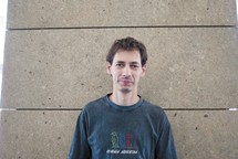 portrait d'Emmanuel, t-shirt bleu foncé, mur brun clair