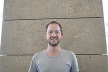 portrait de Sébastien, t-shirt gris, barbe, cheveux courts châtains, sourire, mur brun clair
